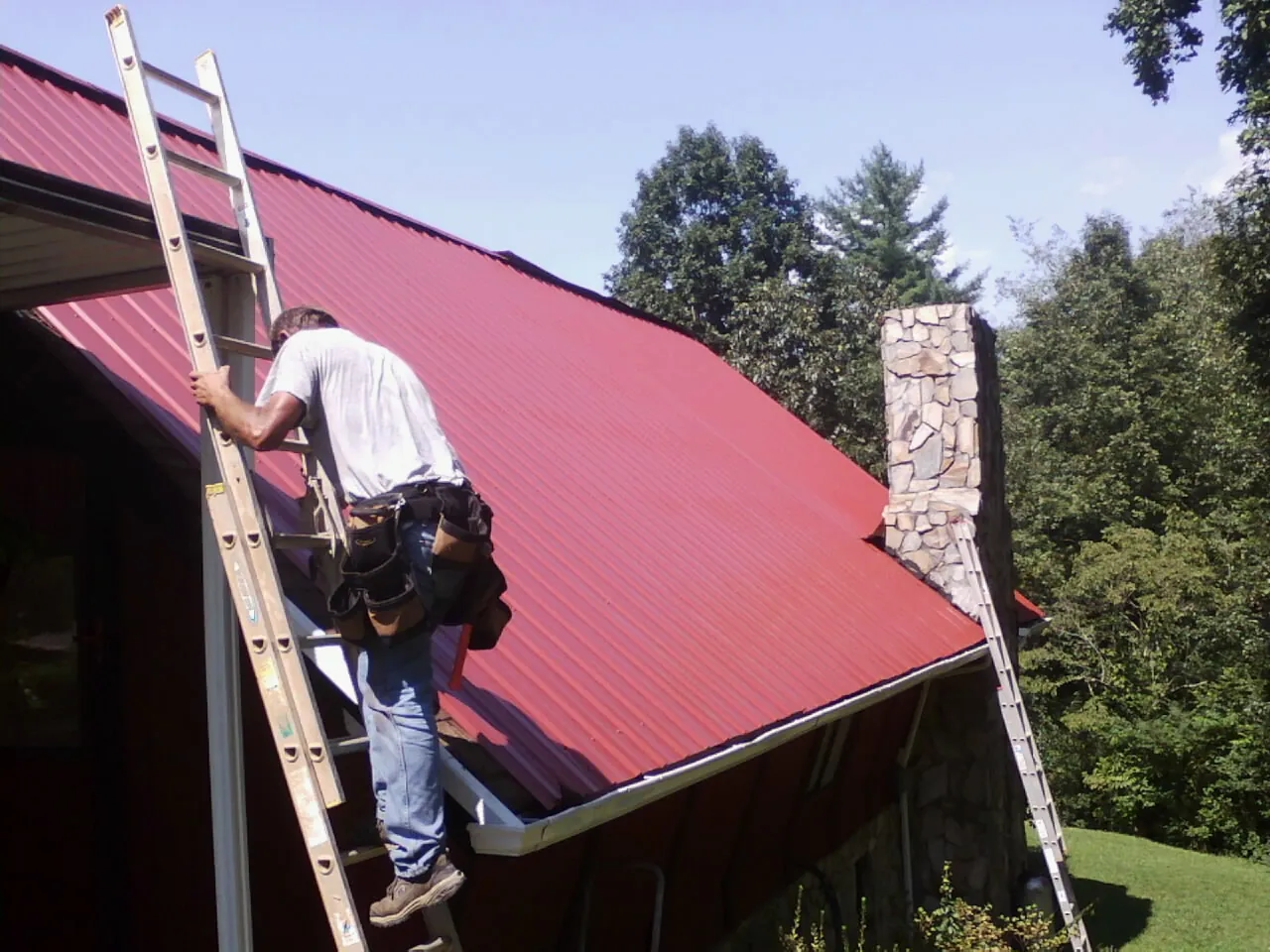 Roofer on a ladder beside a red metal roof and stone chimney — Soffit & Fascia Repair by Roofing Brothers in Walden